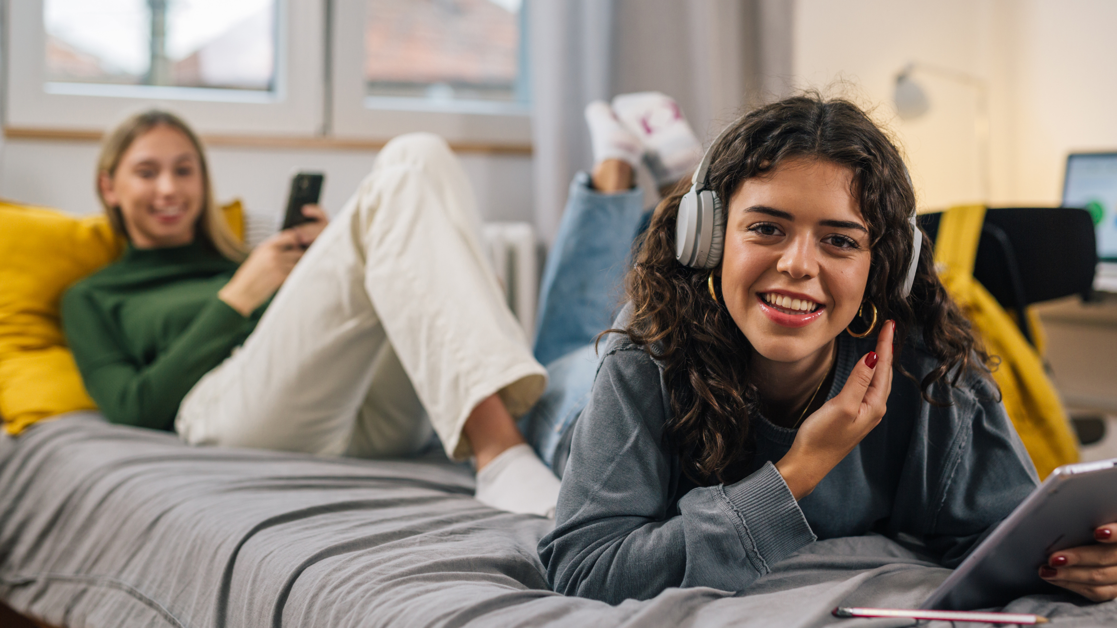 2 women hanging out in their dorm, one is on her phone and the other is smiling staring at the camera with headphones on